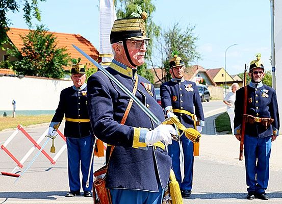 Vier ältere Männer in Uniform stehen auf einer Straße. Der Mann vorne hält ein Schwert und trägt eine Brille. Hinter ihm stehen drei weitere Männer in Uniform. Sie sind wahrscheinlich bei einer Parade. Dahinter befinden sich Häuser, eine Straßenlaterne und ein Auto.