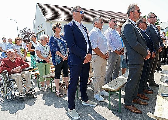 Eine Gruppe von Menschen in Anzügen und Sonnenbrillen steht vor einem Gebäude. Hinter ihnen sitzen mehrere Personen auf Bänken und ein Mann im Rollstuhl.