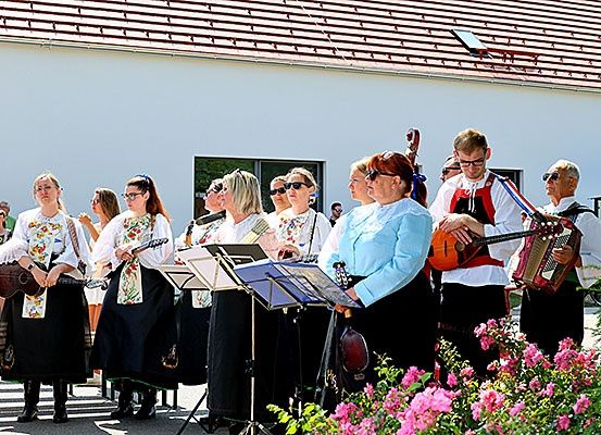Eine Gruppe von Menschen in traditioneller Kleidung steht draußen und spielt verschiedene Instrumente wie Gitarren und Akkordeons. Sie befinden sich vor einem weißen Gebäude mit rotem Dach und einem Fenster.
