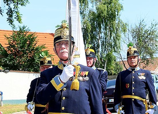 Eine Gruppe von Männern in Uniform, möglicherweise Militär, marschiert bei einer Parade. Sie tragen Mützen, halten Schwerter und folgen einer Fahne. Dahinter sind ein Auto und Bäume zu sehen.