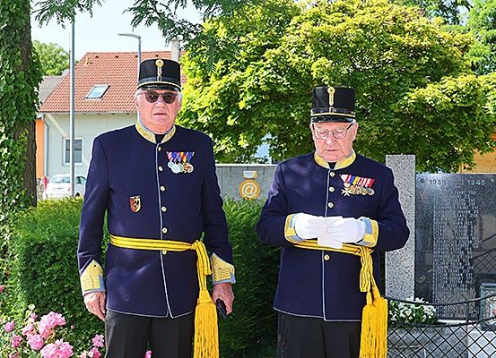 Zwei ältere Männer in Marineuniformen mit Medaillen stehen neben einem Denkmal in einem Garten. Hinter ihnen steht ein Haus und Bäume.