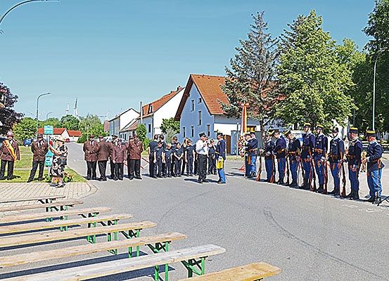 Eine feierliche Veranstaltung auf einem Stadtplatz mit uniformierten Personen, Zuschauern und Holzbänken unter klarem Himmel.