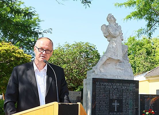 Ein Mann steht an einem Rednerpult und spricht in ein Mikrofon. Hinter ihm steht eine Statue und eine Tafel mit Namen und einem Kreuz. Bäume und ein Gebäude sind im Hintergrund.