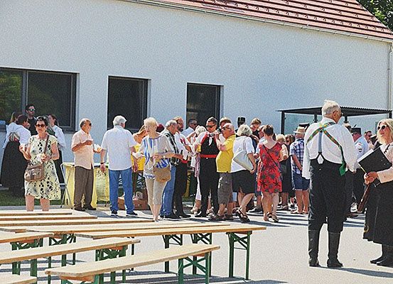 Eine Gruppe von Menschen in traditioneller Kleidung wartet vor einem Gebäude mit Bänken unter einem roten Dach. Im Hintergrund sind Bäume und Fenster zu sehen.
