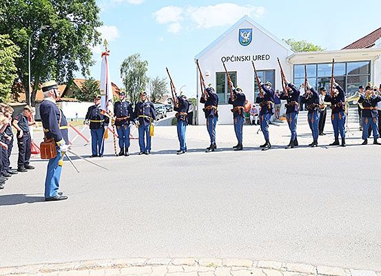 Eine zeremonielle Veranstaltung mit einer Gruppe von Menschen in Uniform, die Gewehre und eine Fahne halten, vor einem Gebäude mit der Aufschrift Opcinski Ured.