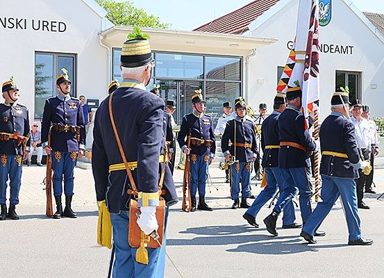 Eine Gruppe uniformierter Personen, wahrscheinlich eine Marschkapelle, zieht in Formation vor einem Gebäude mit der Aufschrift 'Cinki' vorbei.
