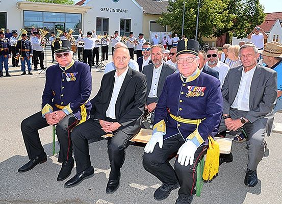 Eine Gruppe von Männern in Uniformen sitzt auf einer Bank draußen, mit einer Band, die hinter ihnen spielt.
