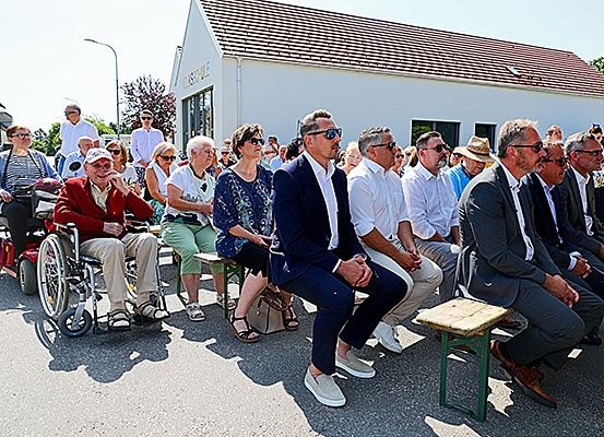 Eine Gruppe von Menschen, darunter ältere Personen, sitzt vor einem Gebäude. Einige tragen Hüte, Brillen und Turnschuhe. Ein Rollstuhl ist vorhanden.