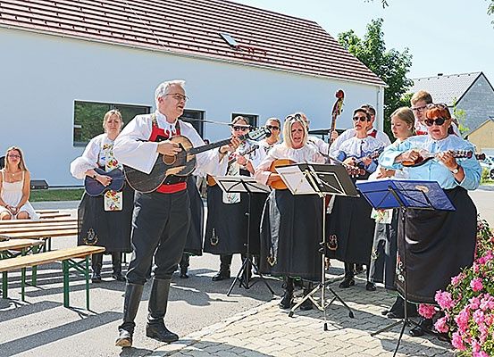 Eine Gruppe von Musikern in traditioneller Kleidung tritt an einem sonnigen Tag vor einem Gebäude auf, wobei eine Frau auf einer Bank im Hintergrund sitzt.