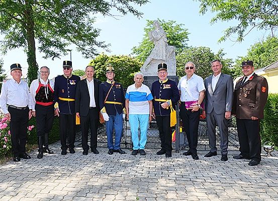 Eine Gruppe von Menschen in formeller Kleidung steht vor einer Statue, einige tragen Hüte und halten Bücher. Die Statue ist von Grünpflanzen umgeben.