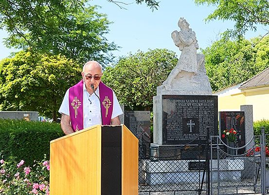Ein Priester in einer violetten Stola steht an einem Rednerpult vor einer Gedenkstatue, umgeben von Blumen und Grün.