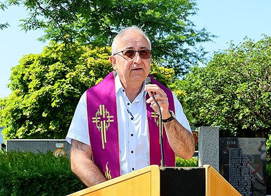 Ein älterer Mann steht an einem Rednerpult, spricht in ein Mikrofon und trägt eine violette Stola mit Kreuzdesign. Die Umgebung ist im Freien, mit Bäumen und einem Gedenkstein hinter ihm.