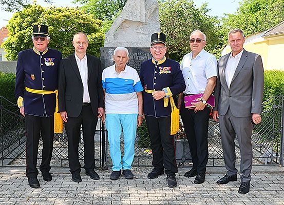 Sechs Männer stehen vor einem Denkmal, tragen verschiedene Uniformen und Schuhe. Der Mann in der Mitte trägt ein weißes und blaues Hemd und blaue Hosen.