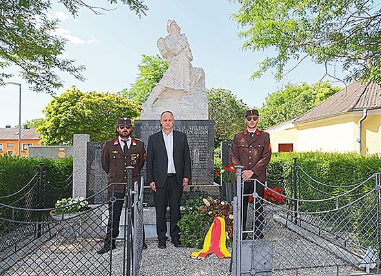 Drei Männer, zwei in Militäruniformen und einer in einem Anzug, stehen vor einer Statue mit Blumen und Flaggen.