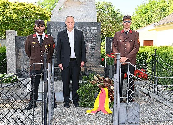 Drei Männer stehen vor einem Denkmal, einer in einem Anzug und zwei in Militäruniformen. Ein Kranz und Blumen sind auf dem Denkmal abgelegt.