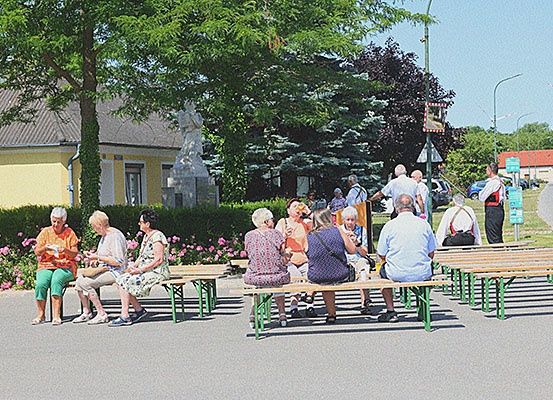Menschen sitzen auf Bänken in einem Außenbereich mit Bäumen und Büschen. Ein Gebäude mit einer Statue ist in der Nähe, und einige Menschen laufen in der Ferne.