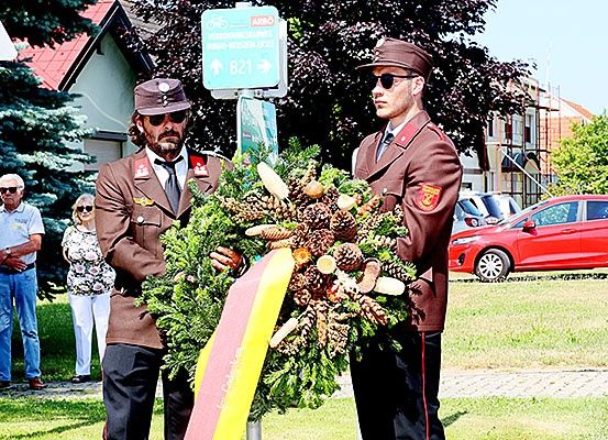 Zwei Männer in Militäruniformen halten eine Girlande im Freien. Hinter ihnen beobachten ein Mann und eine Frau. Ein Straßenschild ist über ihnen. Im Hintergrund ist ein rotes Auto geparkt.