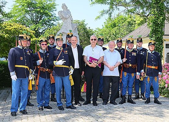 Eine Gruppe von Männern in Uniform steht vor einer Statue, wobei zwei Männer in Zivilkleidung lächeln und ein Buch halten.