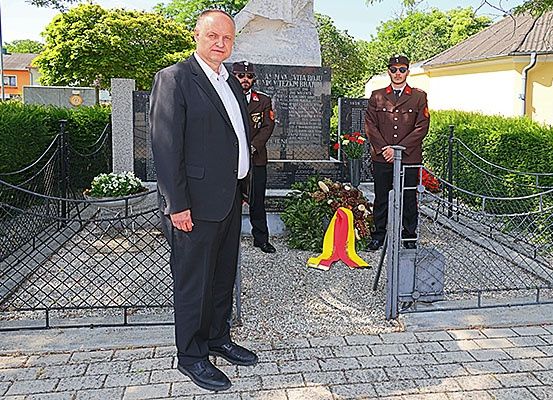 Ein Mann in einem Anzug steht vor einem Denkmal mit zwei uniformierten Offizieren, einer hält einen Kranz, unter einem sonnigen Himmel.