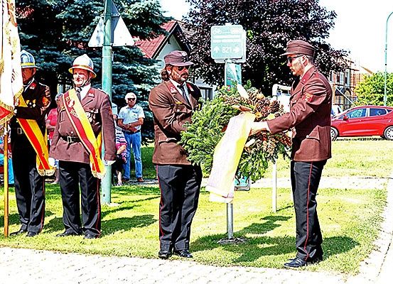 Vier Männer in Militäruniformen stehen auf einem Rasenfeld und halten eine Kranz und eine Flagge. Hinter ihnen schauen Leute zu. Ein rotes Auto ist auf der Seite geparkt.