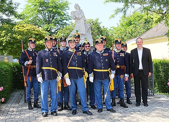 Eine Gruppe von Männern in Uniformen, möglicherweise Militärangehörige, posiert für ein Foto vor einer Statue und einem Haus mit Bäumen und Blumen im Hintergrund.