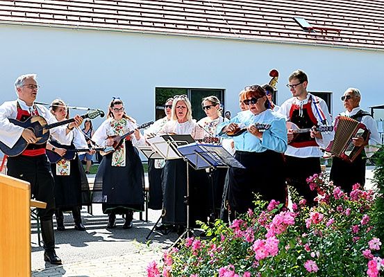 Eine Gruppe von Musikern, einige spielen Gitarren und andere ein Akkordeon, spielen im Freien vor einem Gebäude. Sie sind von Notenblättern und einem Blumenbeet umgeben.
