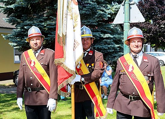 Drei Männer in braunen Uniformen und Helmen halten Fahnen. Sie stehen in einem Park mit Bäumen und einem Gebäude im Hintergrund.