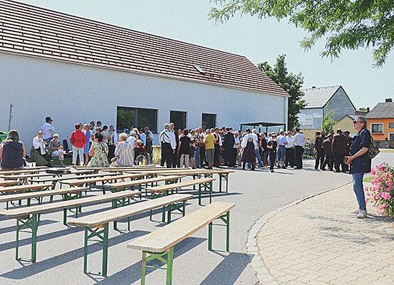 Eine Menschenmenge steht vor einem weißen Gebäude, mit vielen aufgestellten Bänken und Tischen. Die Atmosphäre ist festlich, mit rosa Blumen rechts und Bäumen im Hintergrund.