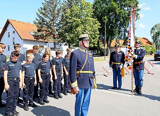 Eine Gruppe von Kindern in Uniformen steht in einer Reihe vor einem älteren Mann in Uniform, der eine Fahne hält. Sie stehen am Straßenrand mit Häusern und Bäumen im Hintergrund.
