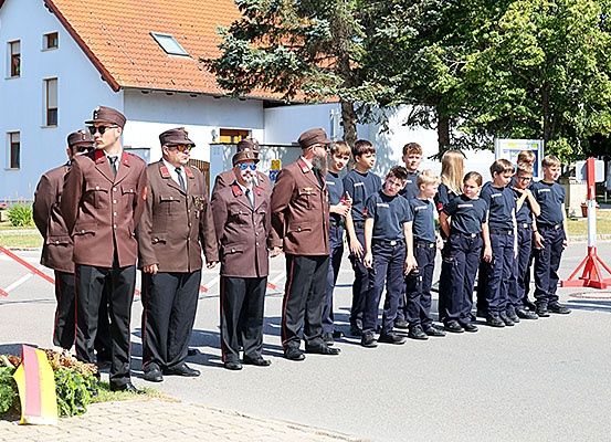 Eine Gruppe von Feuerwehrleuten in Uniformen steht in einer Reihe auf der Straße, einige Kinder in blauen Uniformen in der Nähe, ein Gebäude mit einem roten Dach hinter ihnen und eine Flagge auf der linken Seite.
