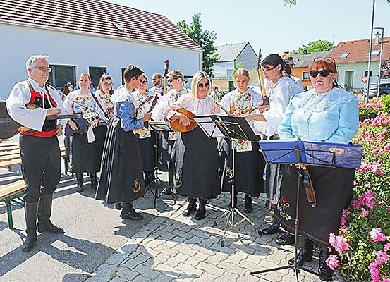 Eine Gruppe von Musikern in traditioneller Kleidung, die verschiedene Instrumente halten, steht vor einem Gebäude mit Häusern und Grünpflanzen im Hintergrund.