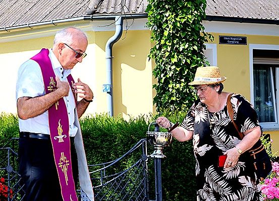 Ein älterer Mann in einer Priesterkleidung und eine Frau mit einer Kerze vor einem Haus mit einem Schild.