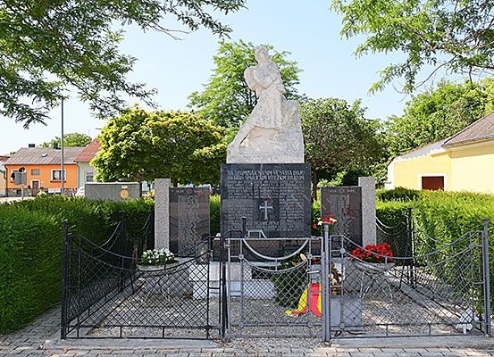 Ein Denkmal in einem Friedhof zeigt eine Statue eines Mannes auf einem Felsen mit einem Kreuz an der Wand. Es ist von Grünpflanzen umgeben, darunter ein Busch und ein Baum. Es gibt zwei Tore, von denen eines eine Blumenanordnung hat.