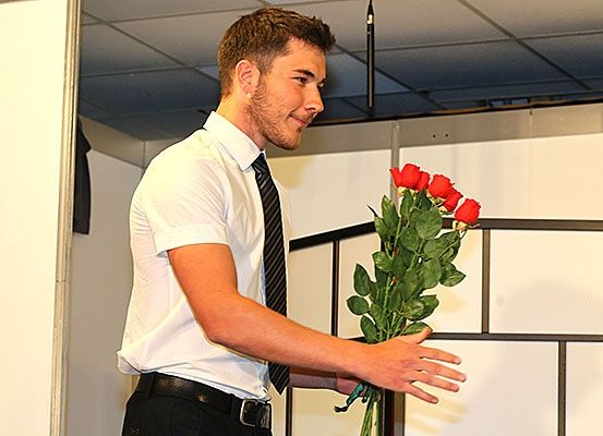 Ein Mann in einem weißen Hemd und einer schwarzen Krawatte hält einen Strauß roter Rosen und steht in einem Raum mit weißen Wänden und einer offenen Tür.