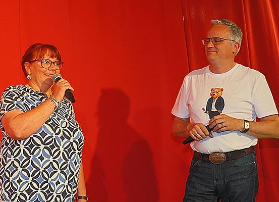 Eine ältere Frau und ein Mann stehen vor einem roten Vorhang, wahrscheinlich auf einer Bühne. Die Frau hält ein Mikrofon, während der Mann ein Mikrofonstativ hält.