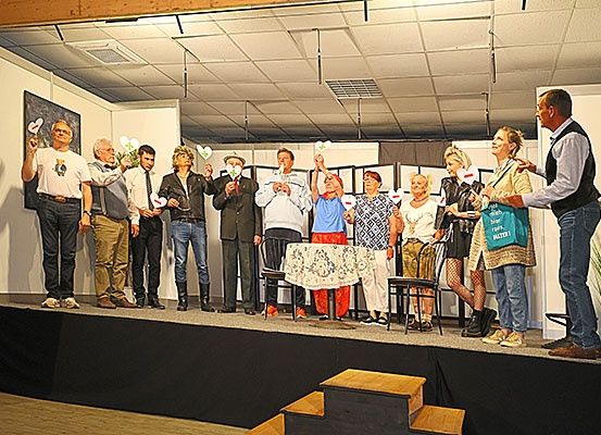 Eine Gruppe Erwachsener steht auf einer Bühne, wahrscheinlich bei einer Aufführung oder Veranstaltung, und hält herzförmige Gegenstände. Sie sind lässig gekleidet, und einige haben Krawatten.