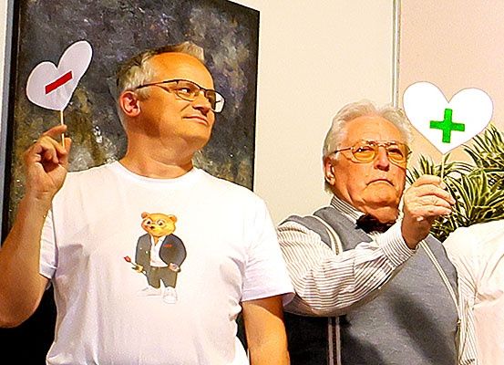 Zwei Männer, einer mit einem Herz mit Minuszeichen, der andere mit einem Herz mit Pluszeichen, posieren für ein Foto vor einem Gemälde.