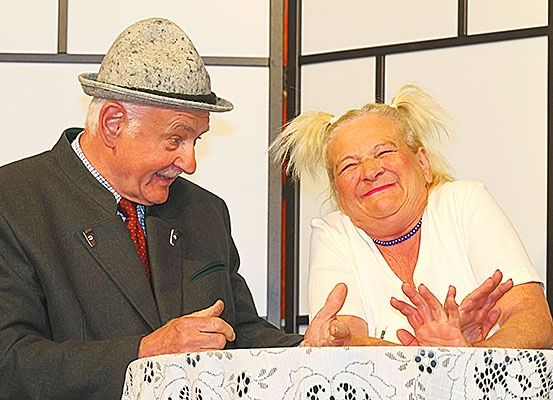 Ein älterer Mann und eine ältere Frau sitzen an einem Tisch, lächeln und gestikulieren. Der Mann trägt einen grauen Hut und Anzug, die Frau ein weißes Hemd und eine Halskette.