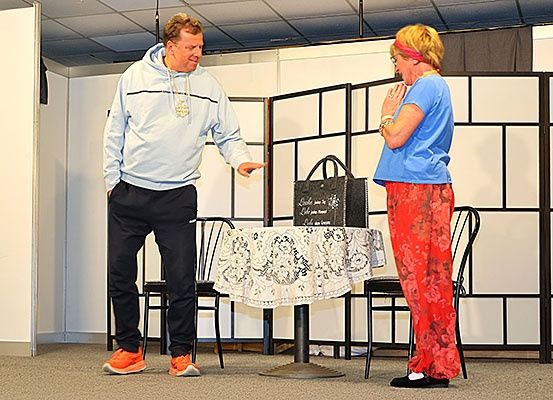 Ein Mann und eine Frau stehen neben einem Tisch, anscheinend mitten in einem Gespräch. Der Mann trägt eine Kapuzenjacke und Turnschuhe, während die Frau ein blaues T-Shirt und rote Hosen trägt. Hinter ihnen steht ein Tisch, der mit einer Tischdecke versehen ist, und eine Handtasche ruht darauf.