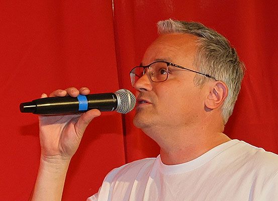 Ein Mann mit grauem Haar spricht in ein Mikrofon, das er mit der rechten Hand hält. Er trägt eine Brille und ein weißes T-Shirt. Hinter ihm befindet sich ein roter Vorhang.