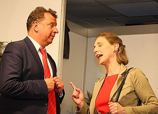Ein Mann in einem Anzug und eine Frau in einem roten Oberteil führen ein Gespräch. Der Mann lächelt und zeigt auf die Frau. Die Frau lacht und zeigt zurück.