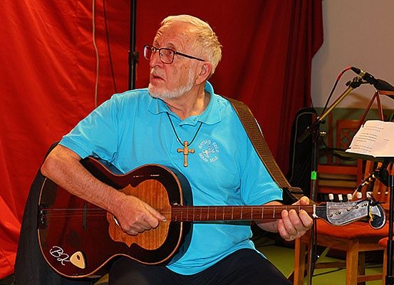Ein älterer Mann in einem blauen Hemd und mit Brille spielt eine Gitarre auf der Bühne mit einem roten Vorhang im Hintergrund.