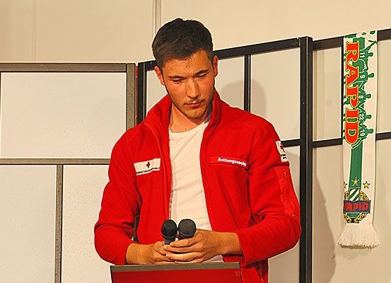 Ein Mann in einem roten Jacken steht an einem Podium mit zwei Mikrofonen und spricht in sie. Hinter ihm ist eine weiße Wand und ein Banner mit grünem Text zu sehen.