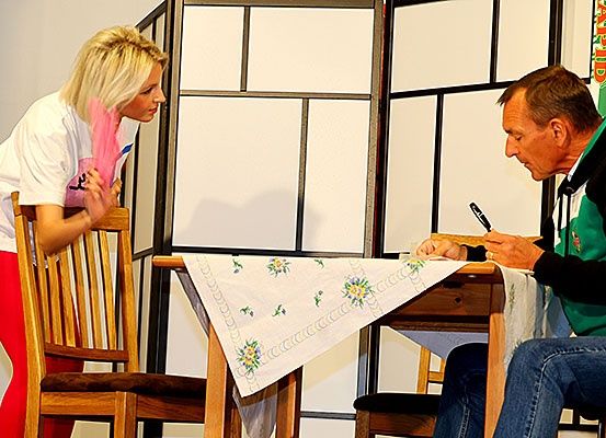 Eine Frau steht hinter einem Mann, der an einem Tisch sitzt, und hält eine Feder in der Hand. Der Mann schreibt mit einem Stift auf einem Papier und konzentriert sich darauf.