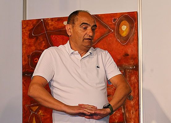 Ein älterer Mann steht mit gefalteten Händen vor einem großen, abstrakten Gemälde. Er trägt ein weißes Poloshirt und eine grüne Uhr.