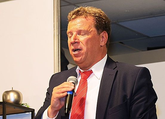 Ein Mann in Anzug und roter Krawatte hält ein Mikrofon und spricht hinein, möglicherweise bei einer Veranstaltung oder Präsentation.