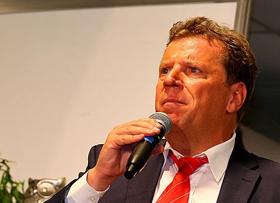 Ein Mann in Anzug und roter Krawatte spricht in ein Mikrofon. Er hat kurze braune Haare.