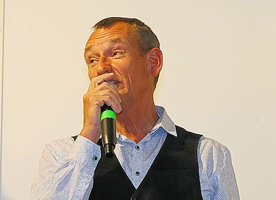 Ein Mann in einem blauen Hemd und schwarzem Weste hält ein Mikrofon an seinen Mund, wahrscheinlich bei einer Veranstaltung auftretend oder sprechend.