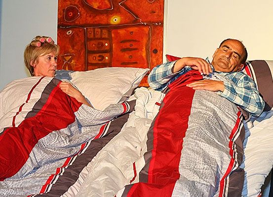 Ein Mann und eine Frau liegen auf einem Bett mit roten und grauen Bezügen, beide ruhen, während ein Gemälde an der Wand hinter ihnen hängt.