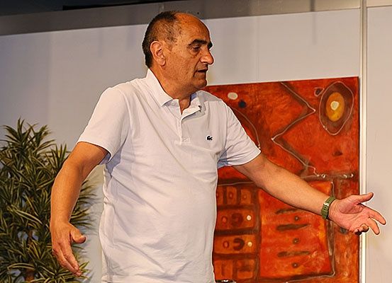 Ein Mann steht drinnen und gestikuliert mit den Händen. Er trägt ein weißes Poloshirt. Hinter ihm befindet sich ein großes abstraktes Gemälde an der Wand.